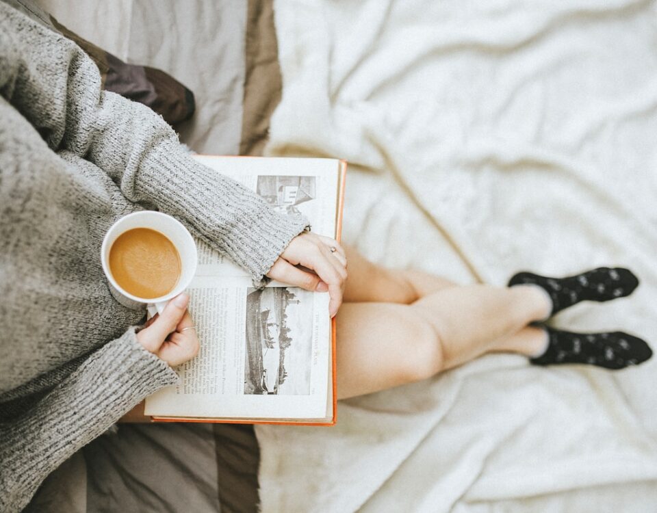 Práticas de autocuidado para evitar o burnout diário woman holding a cup of coffee at right hand and reading book on her lap while holding it open with her left hand in a well-lit room