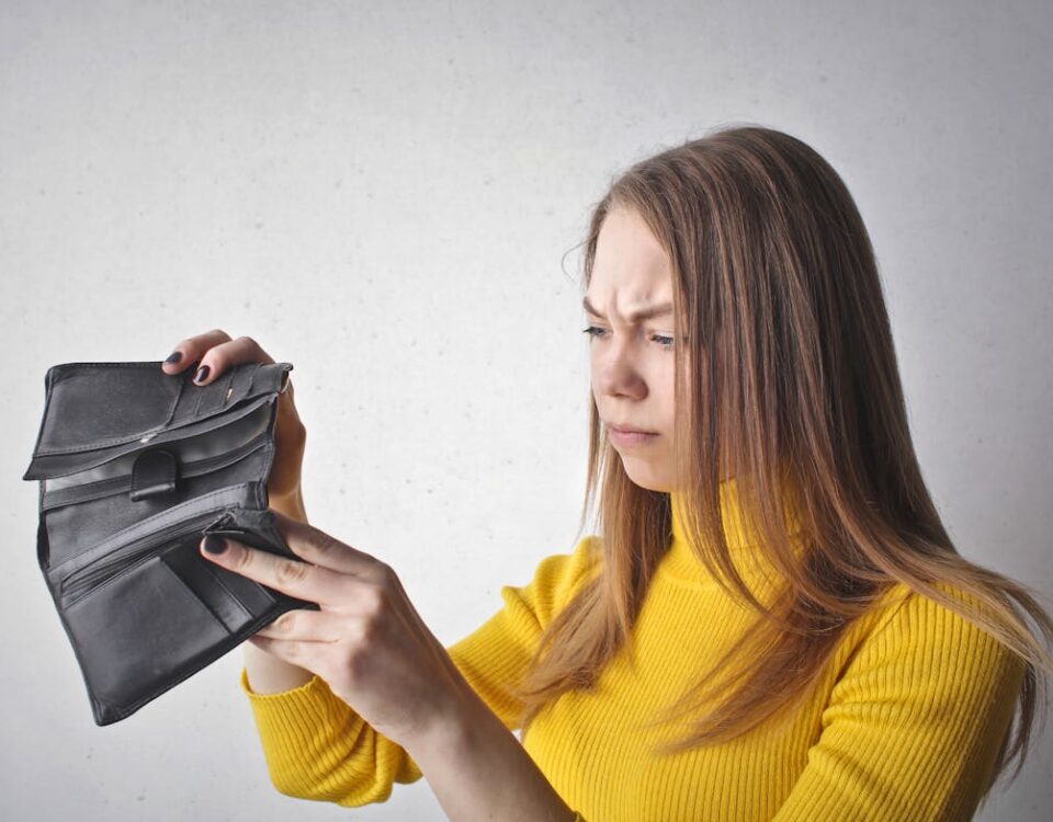 Como o estresse financeiro afeta seu bem-estar Young woman in a yellow top looking at her empty wallet with a frustrated expression.