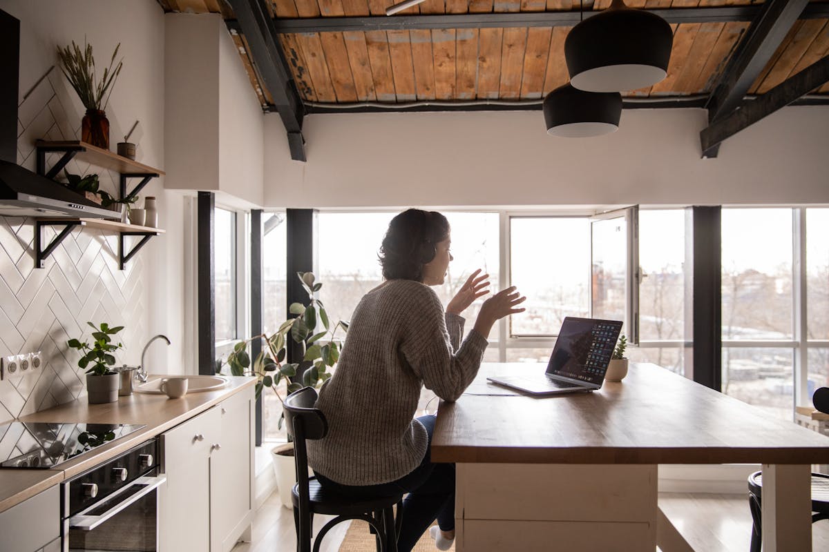 Vantagens práticas dos robôs no atendimento digital Back view of young female expressively talking via laptop while sitting at wooden table in spacious kitchen
