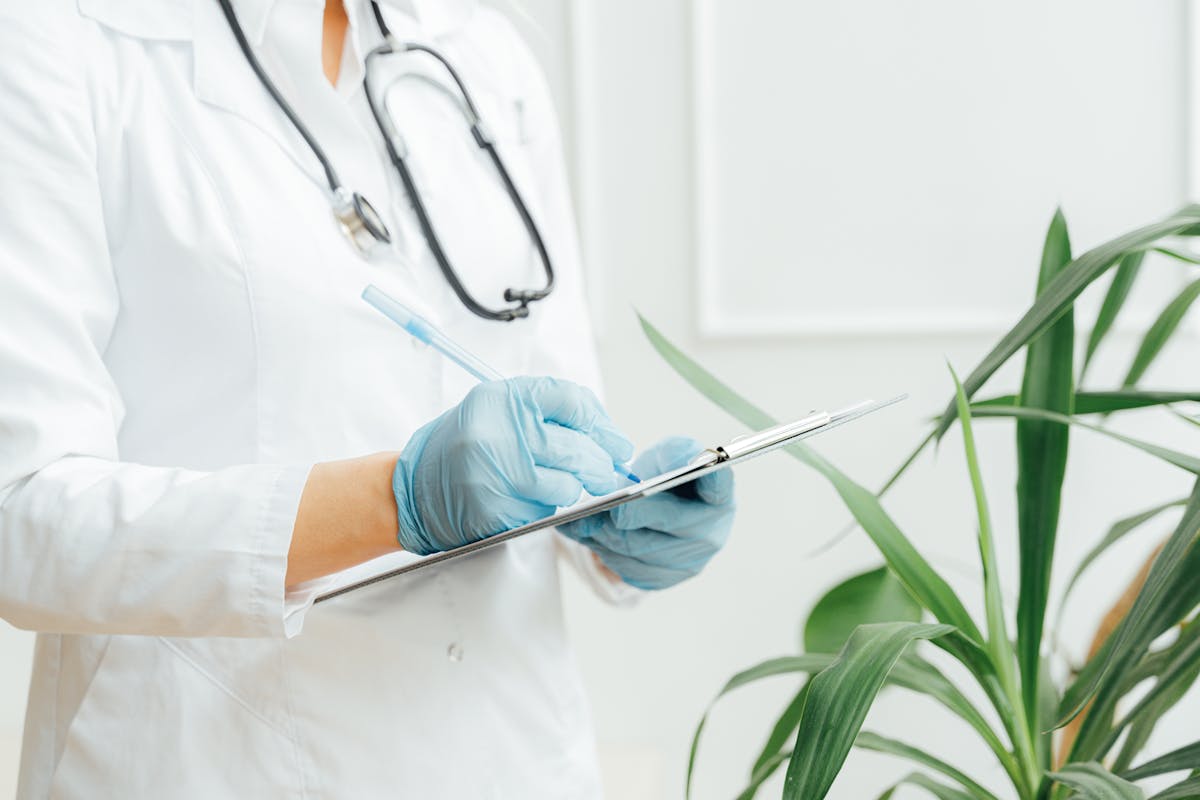 Luvas Descartáveis: Quando Trocar e Qual o Impacto na Segurança do Paciente A healthcare professional wearing gloves takes notes on a clipboard with a stethoscope around their neck.