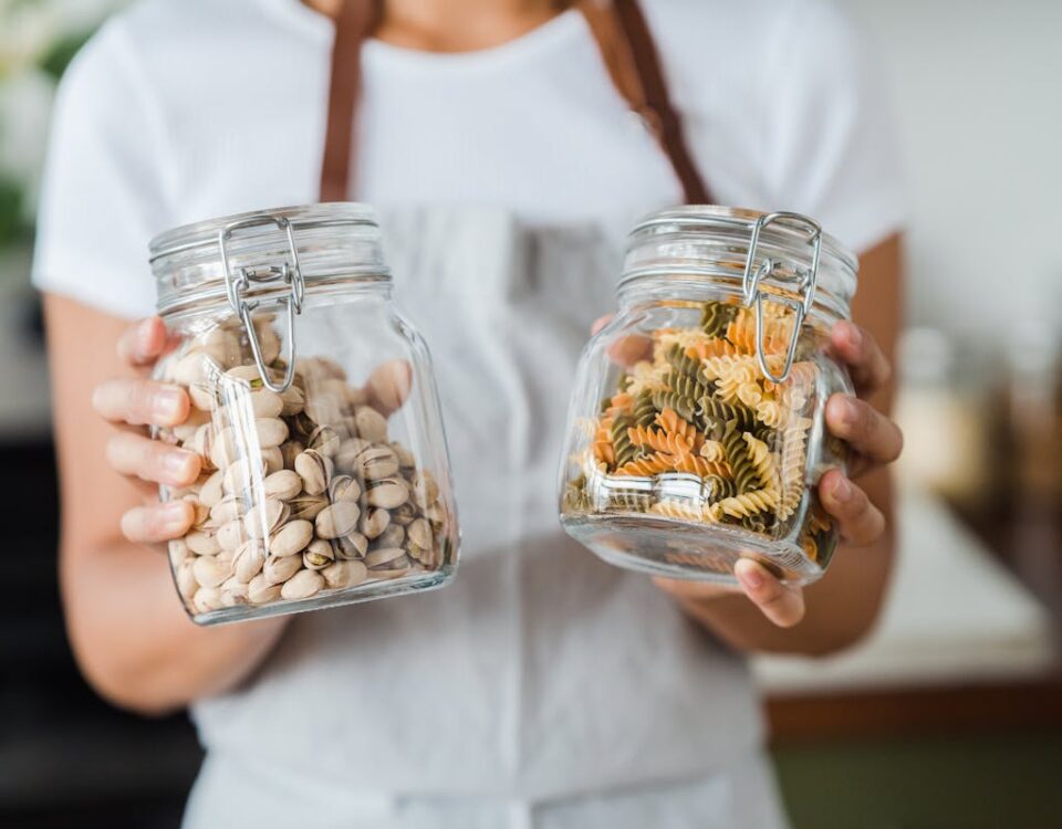 Hands holding glass jars filled with pistachios and colorful spiral pasta indoors.