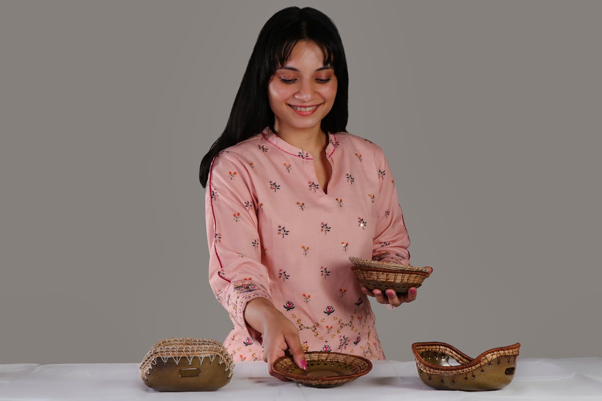 A smiling woman in a pink floral top showcases handcrafted pottery pieces indoors.