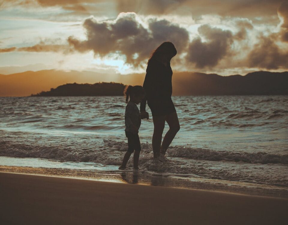 Benefícios de morar perto da natureza A mother and child walk along a Florianópolis beach at sunset, creating a serene, backlit silhouette against the ocean waves.