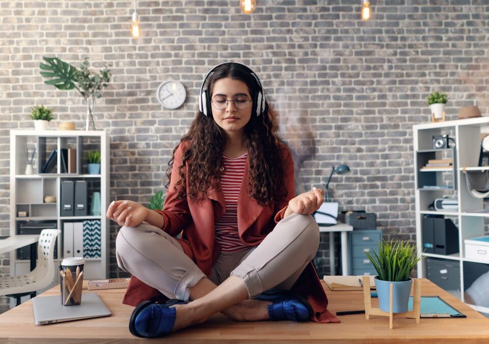 Como manter o equilíbrio na rotina de auxiliar administrativo Young woman meditating with headphones in a modern office setting, practicing mindfulness.