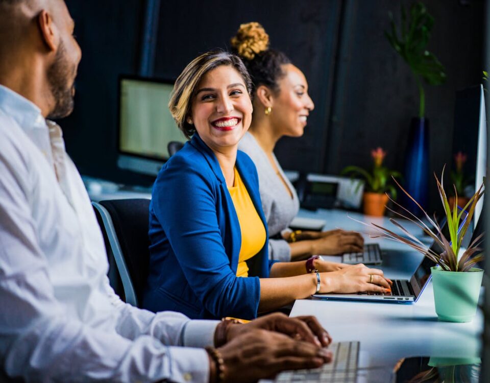 Automação comercial e qualidade de vida no trabalho Three diverse professionals working and smiling at office desks, fostering teamwork and collaboration.