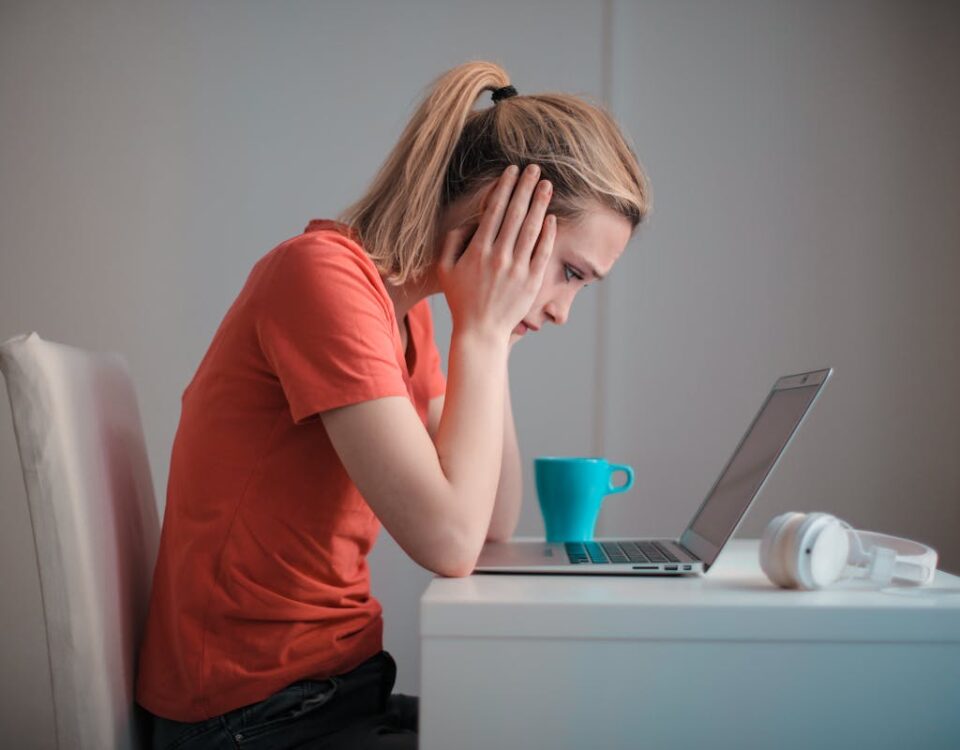 A ansiedade antes da prova de direção: Como lidar? A woman sits indoors at a table, looking concerned while using a laptop with headphones and a coffee mug nearby.