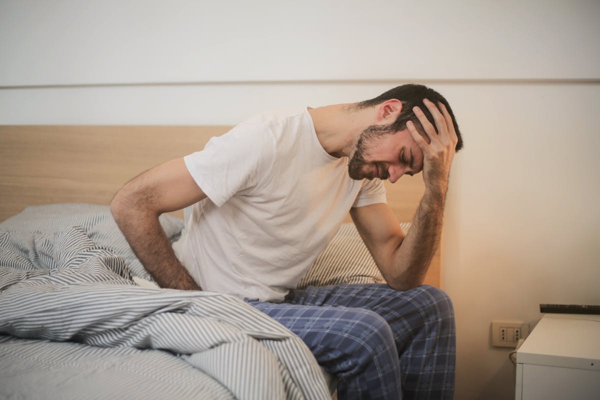 Estilo de vida que minimiza crises de doenças inflamatórias A young man in pajamas holding his head, sitting on a bed, appears to be experiencing a headache.
