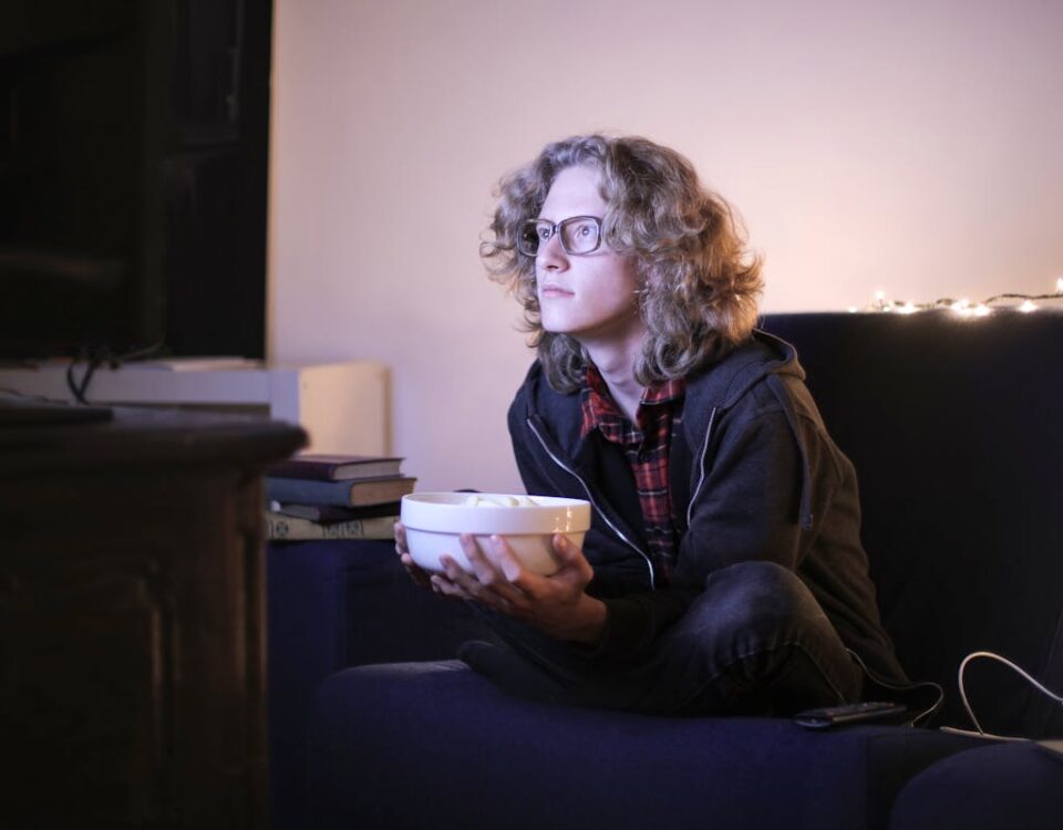 O impacto de assistir filmes no relaxamento diário Young man with glasses watching television with focus while holding a snack bowl in a cozy living room setting.