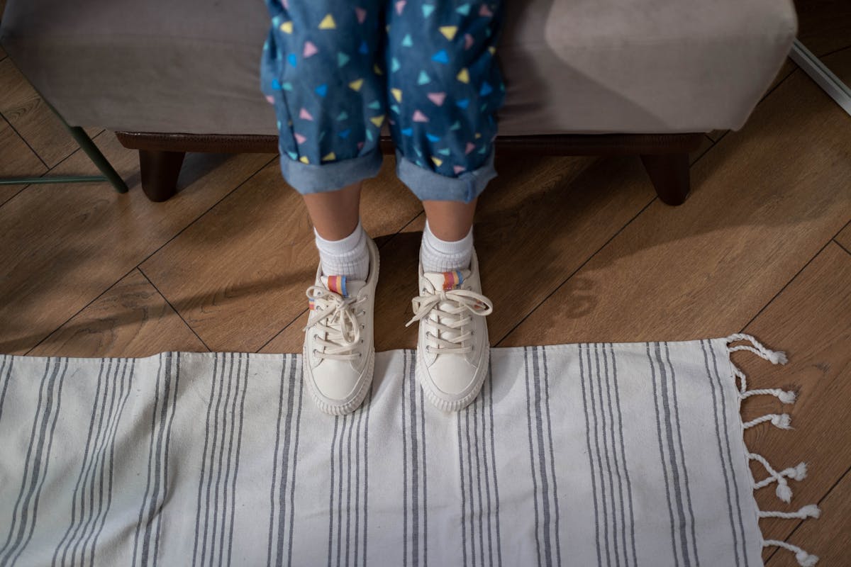 Como calçados adequados impactam a saúde das crianças Child standing on a rug wearing patterned pants and sneakers in a cozy indoor setting.
