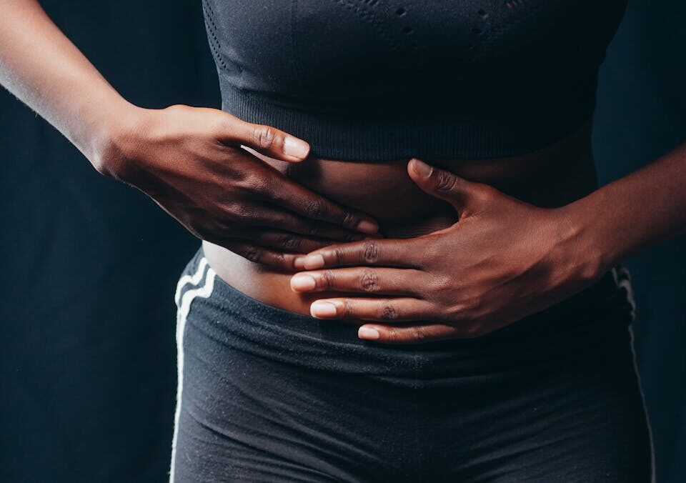 Estilo de vida que minimiza crises de doenças inflamatórias Close-up of a person pressing hands on abdomen, indicating discomfort or pain, against a black background.