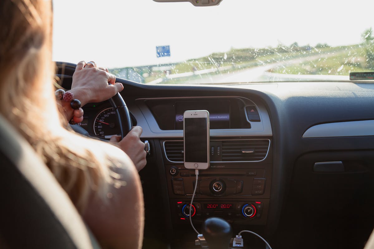 Dicas de alongamento para quem dirige por muitas horas View from behind a woman driving a car on a sunny day with a phone mounted on the dashboard.