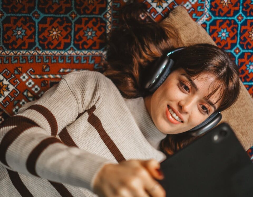 O impacto do áudio no bem-estar diário Brunette woman in turtleneck lying on patterned rug with headphones and tablet.