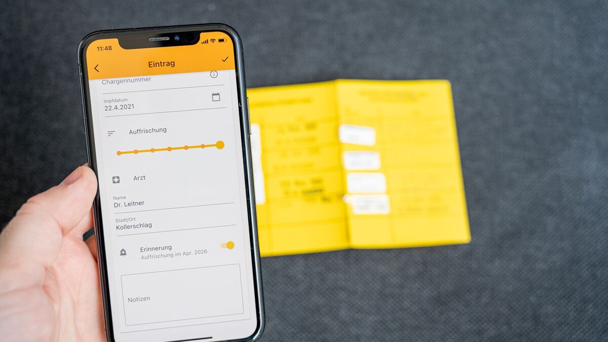 Dia Nacional da Vacinação: como checar sua caderneta e o que atualizar Mão segurando celular com aplicativo de registro de vacinas ao lado de uma caderneta de vacinação amarela