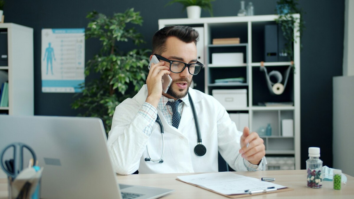 Médico de jaleco falando ao telefone em seu consultório, com notebook e estetoscópio sobre a mesa