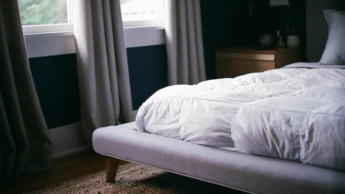 O que o descanso semanal faz pelo corpo de quem trabalha muito? Cama com roupa de cama clara ao lado de uma janela com cortinas